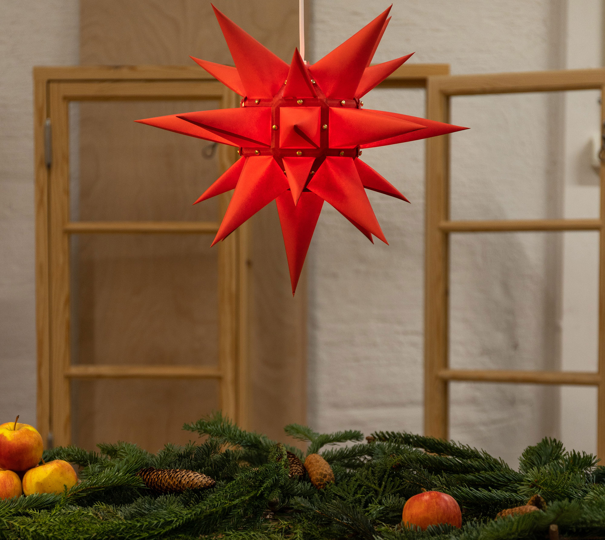 Herrnhuterstjerne i r&oslash;d som julepynt, h&aelig;ngende over gran, kogler og &aelig;bler i klassisk juledekoration.