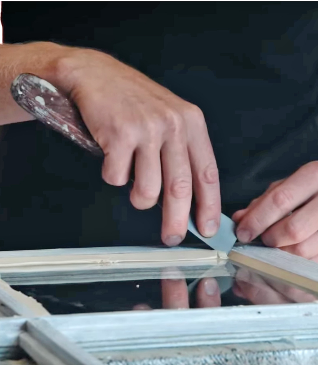 To h&aelig;nder, der kitter et sprossevindue med en kitkniv.