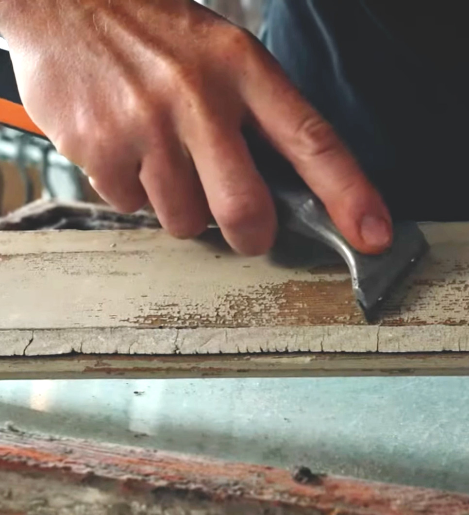 En h&aring;nd skraber gammel maling af en vinduesramme.