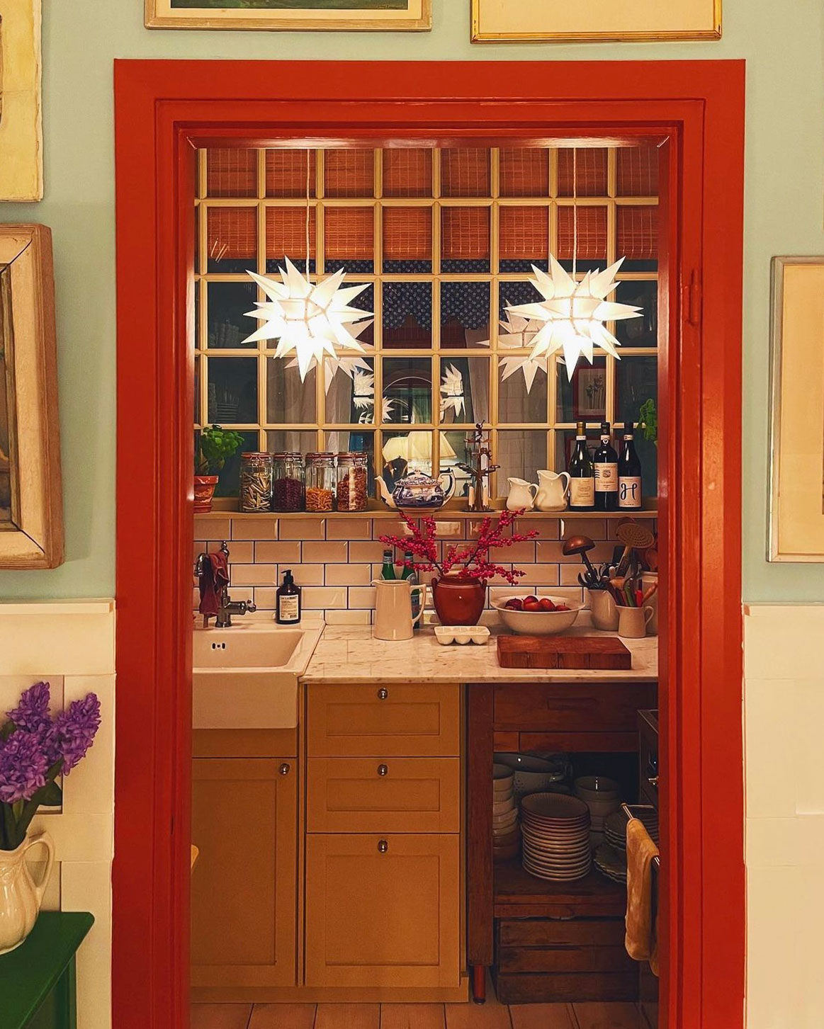 To Herrnhuterstjerner som julestjerner i k&oslash;kkenvindue, set gennem r&oslash;dt d&oslash;rkarme og klassisk indretning.